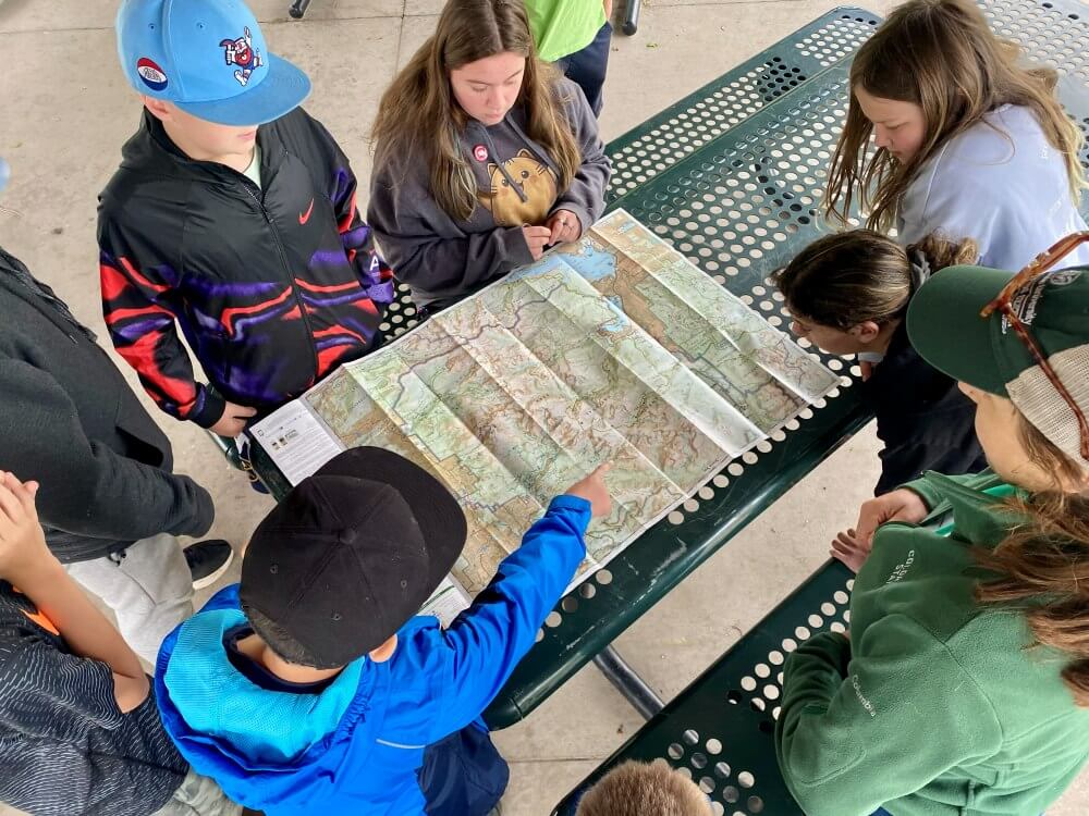 A group of youth gather around a picnic table studying a large folded map, with one person pointing to a specific location during an outdoor activity.