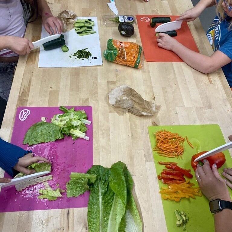 Participants chop vegetables on colorful cutting boards at a table, preparing lettuce, peppers, cucumbers, and other fresh ingredients during a hands‑on food activity.