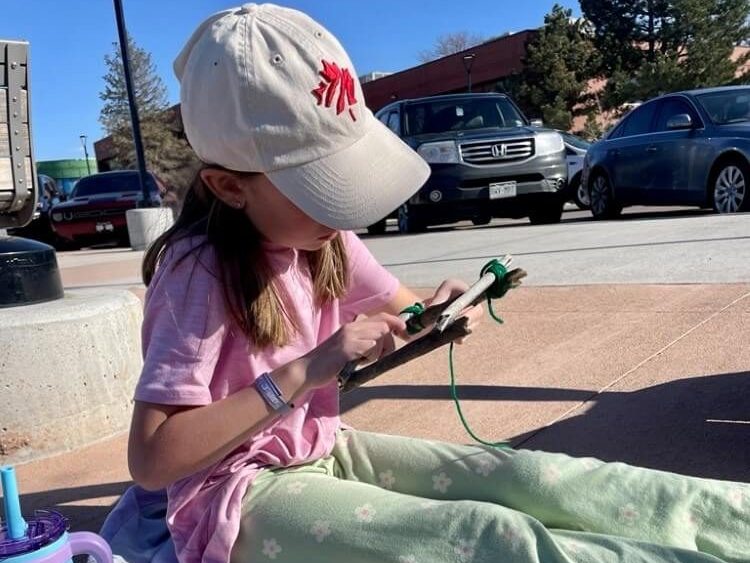 A youth sits on a blanket outdoors weaving green yarn across a small wooden frame, with crafting materials placed nearby.