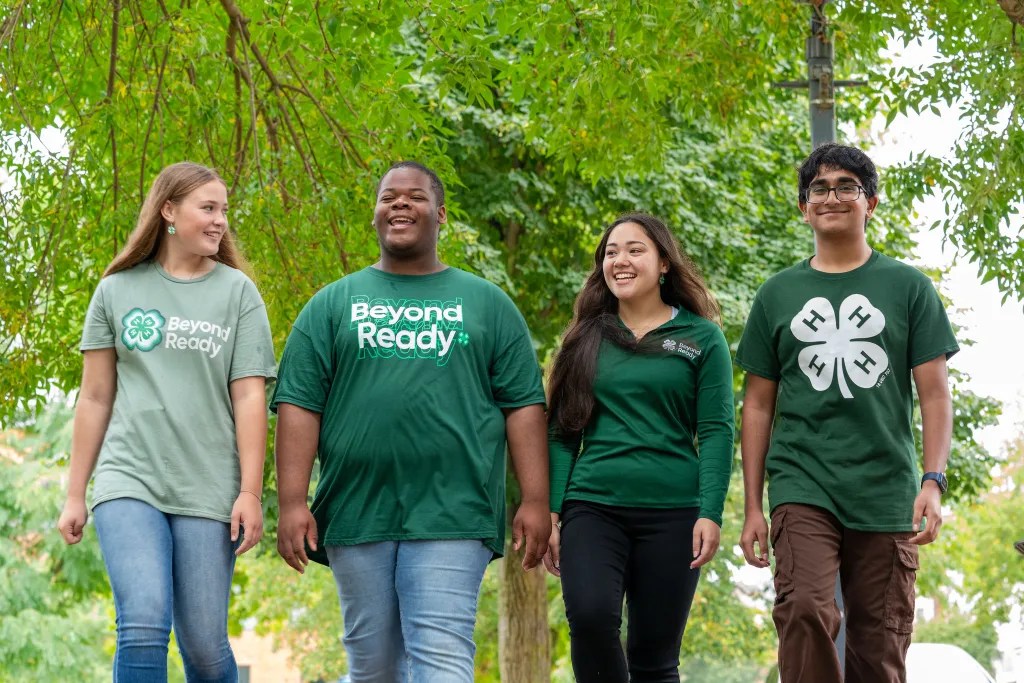 Four youth wearing 4-H shirts walk outside under trees.