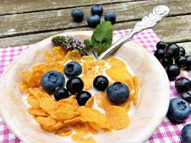 A bowl of cereal topped with blueberries sits on a table, with extra blueberries scattered nearby.