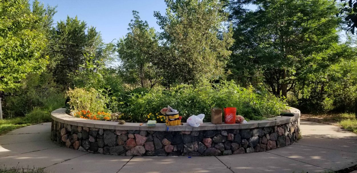 A circular raised garden bed bordered by a stone wall contains dense green plants and flowers, with gardening tools and containers placed along the edge and trees surrounding the area.