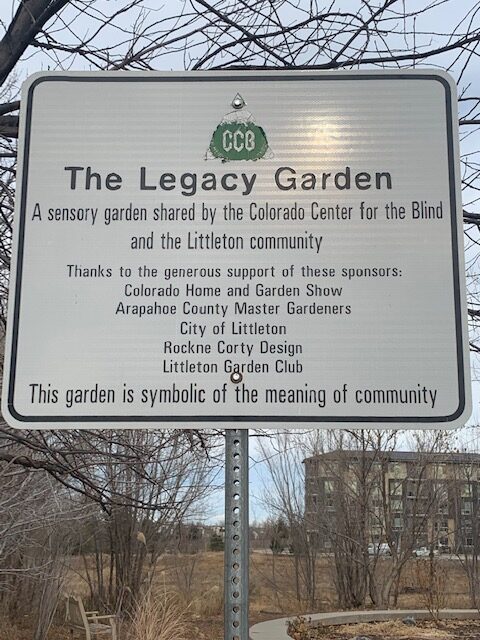 A sign titled “The Legacy Garden” describes a sensory garden supported by community sponsors, mounted on a post in an outdoor area.