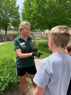 An instructor holds up a plant while speaking to two youth standing nearby in a garden area with raised beds and trees in the background.