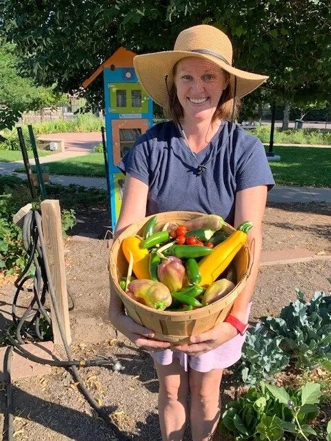 A person stands in a garden holding a basket filled with fresh vegetables, including squash, peppers, and tomatoes.