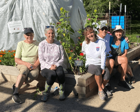 Five volunteers sit on the edge of a raised garden bed surrounded by flowers and greenery in an outdoor garden space.