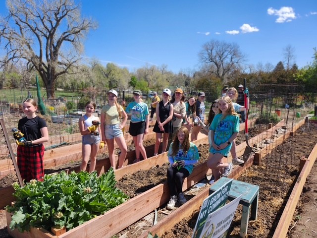 A group of youth stand together in raised garden beds, each holding a plant or tool in an outdoor garden area.