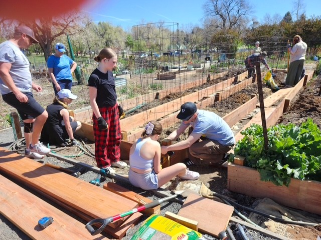 A group of youth and adults work together building and planting raised garden beds, with tools and materials spread around the area.