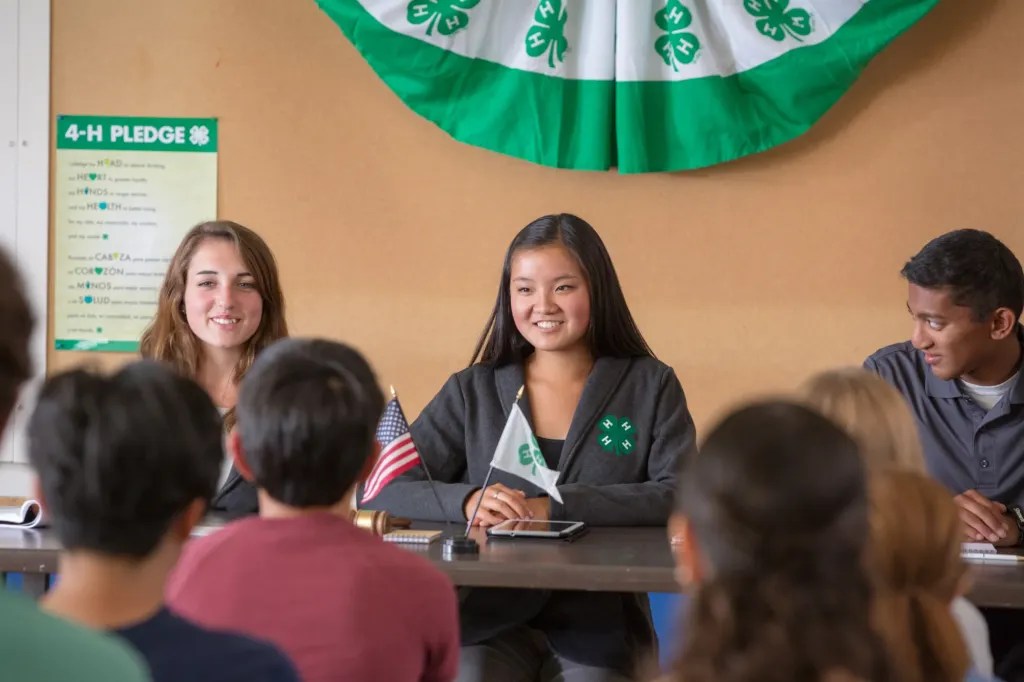 Three individuals are seats and standing behind a table in a meeting room decorated with 4-H banners and flags. The table has small American and 4-H flags, a gavel, and notebooks.