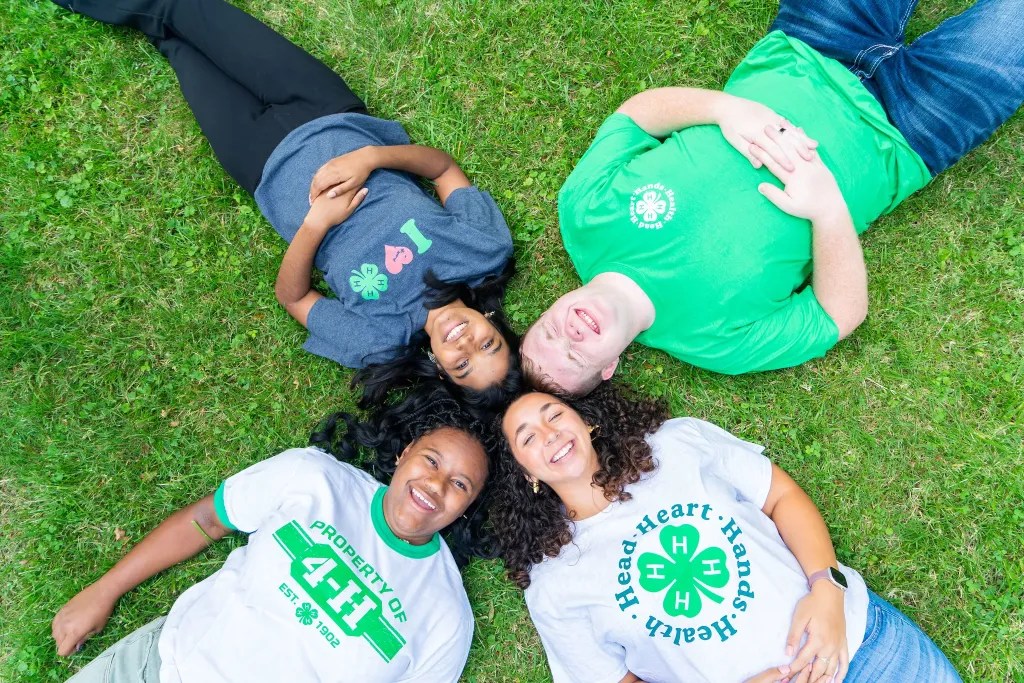 Four 4-H youth members laying on the grass smiling with their heads positioned together in a circle.