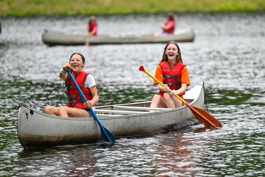 Two girls wearing life jackets paddle a canoe on a lake, smiling and laughing as they row together.