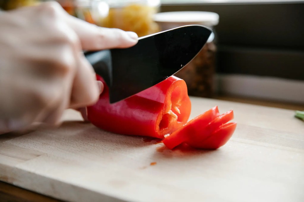 Close-up of a hand slicing a red bell pepper on a wooden cutting board with a black kitchen knife.