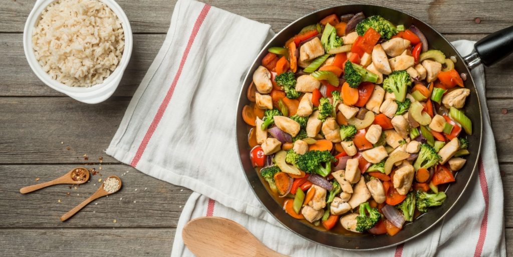 A pan of colorful chicken and vegetable stir-fry sits on a wooden table with a red-striped white towel beneath it.