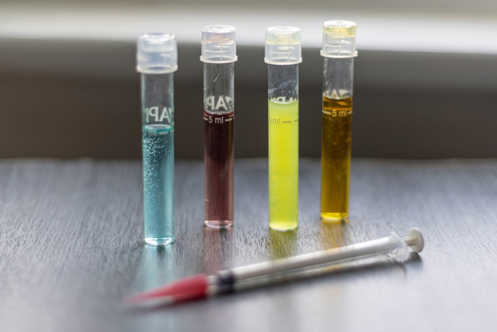 Four small clear vials filled with blue, red, yellow, and amber liquids stand upright on a tabletop, with a syringe placed in front of them.