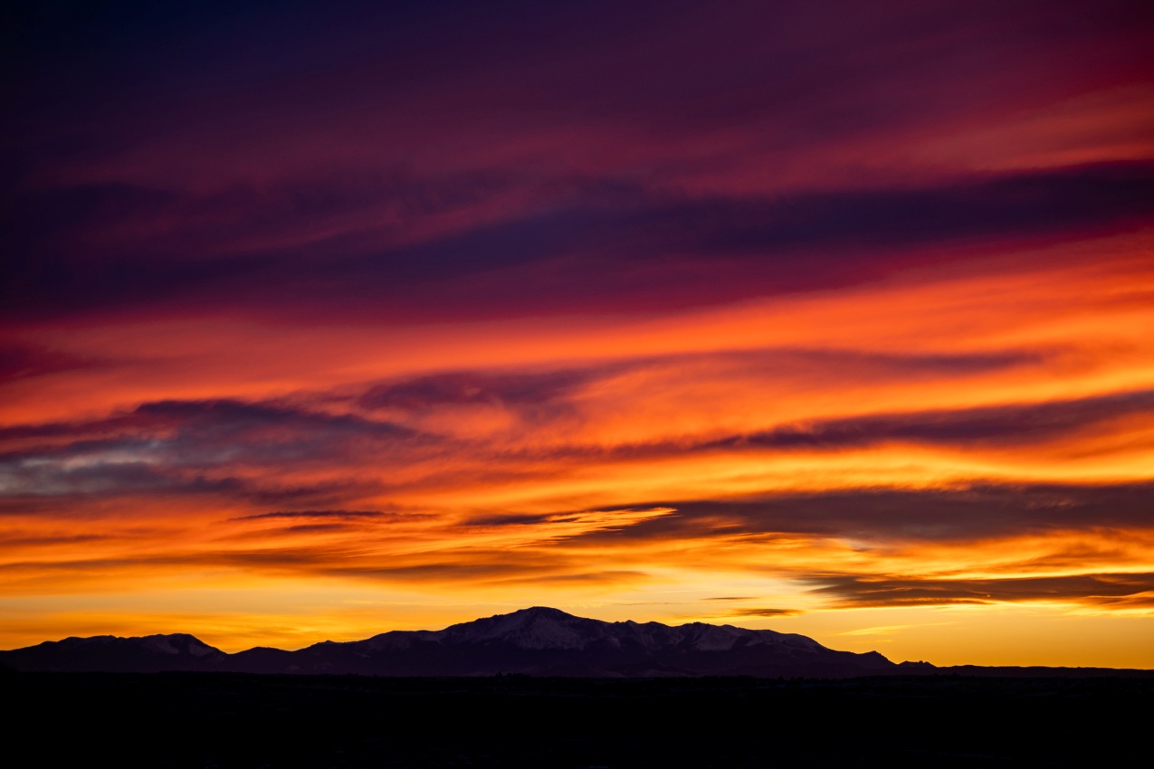 A sunset fills the sky with deep red, orange, and purple clouds above a dark mountain silhouette and shadowed plains below.