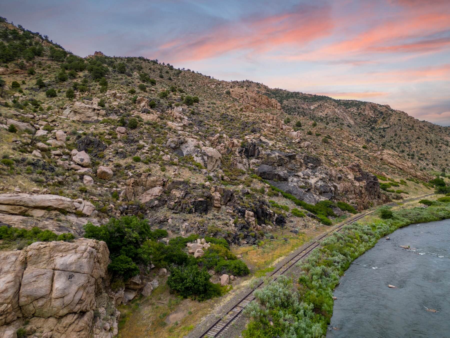 Rocky canyon landscape with a river flowing alongside railroad tracks, with hills and scattered vegetation under a colorful evening sky.