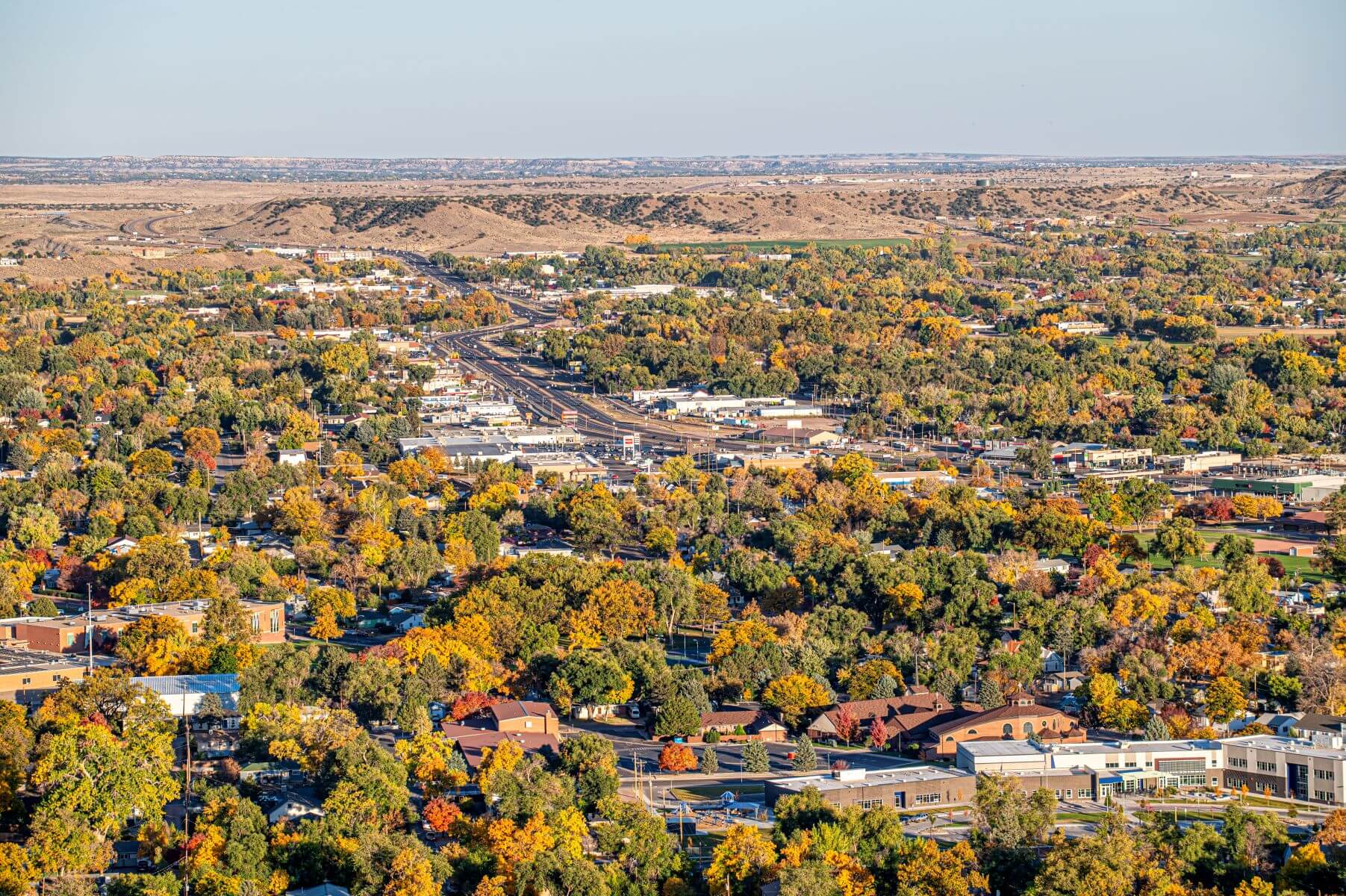 Aerial view of a town with tree‑lined streets, buildings, and a central roadway, surrounded by rolling hills and autumn foliage.