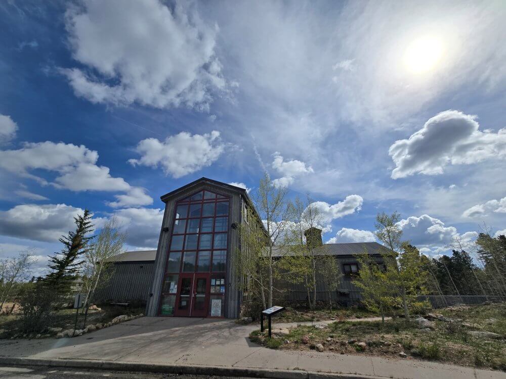 A tall‑fronted building with large windows stands beneath a dramatic sky filled with scattered clouds and bright sun, surrounded by young trees and a paved walkway.