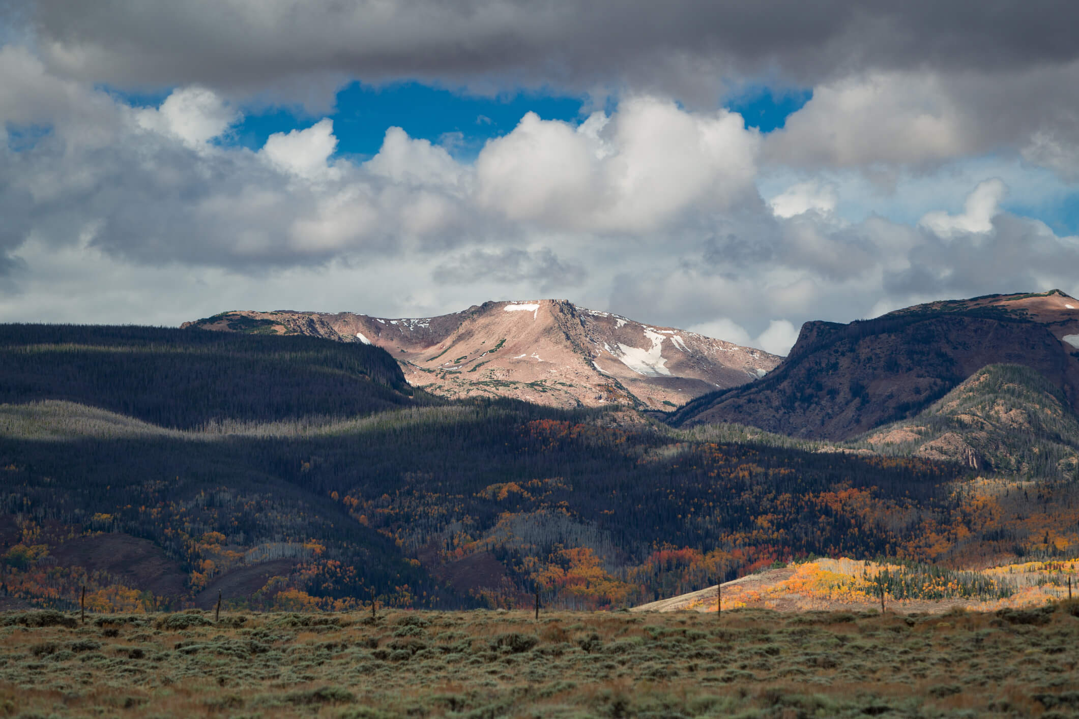 Wide view of a mountain range with rocky peaks and patches of snow above dense evergreen and autumn‑colored forests, under a sky filled with large, layered clouds.