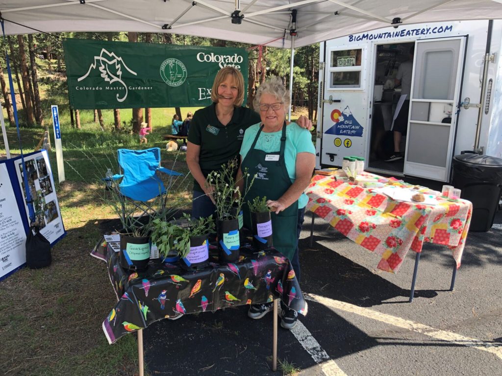 Two Colorado Master Gardener volunteers stand behind a table of labeled plant starts under a canopy tent at an outdoor event, with informational banners and a nearby food truck in the background.
