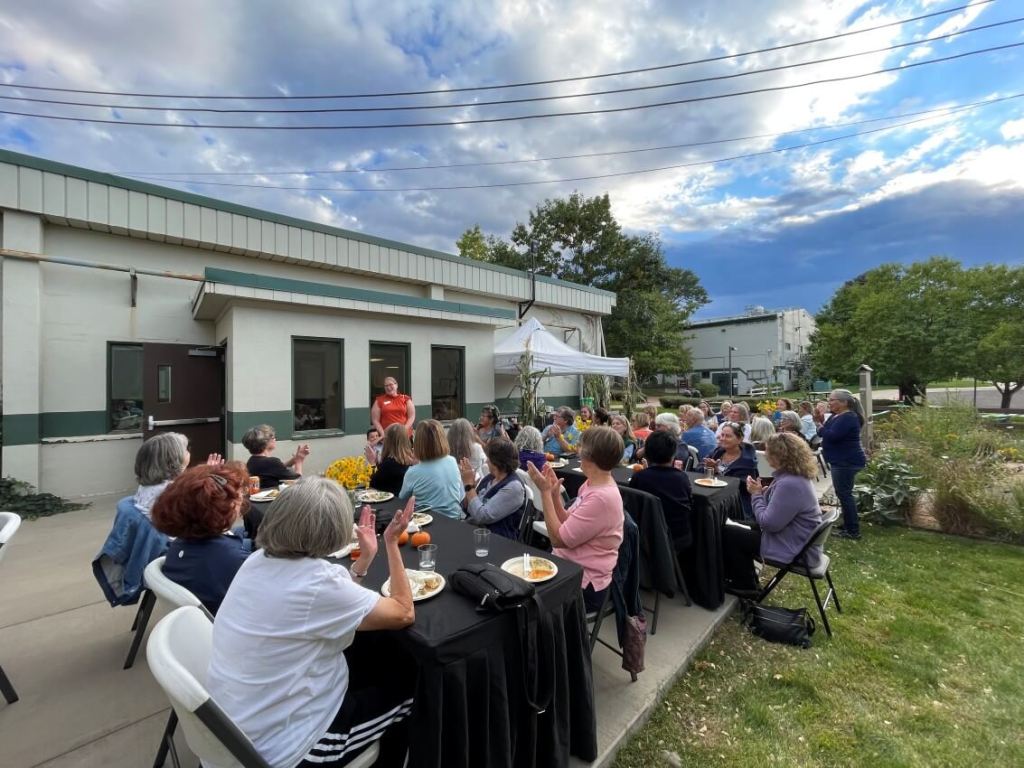 A group of people seated at outdoor tables for a gathering or presentation, with one person standing and speaking near a building on a partly cloudy day.