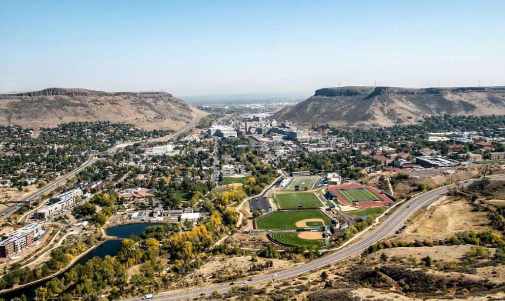 A wide aerial view of the city of Golden, Colorado, showing residential neighborhoods, athletic fields, and downtown buildings nestled between two flat‑topped mesas under a clear sky.