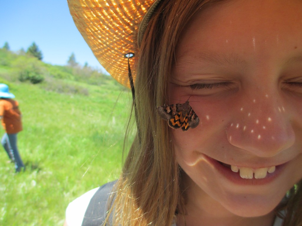 A child smiles at the camera with a butterfly on her cheek.