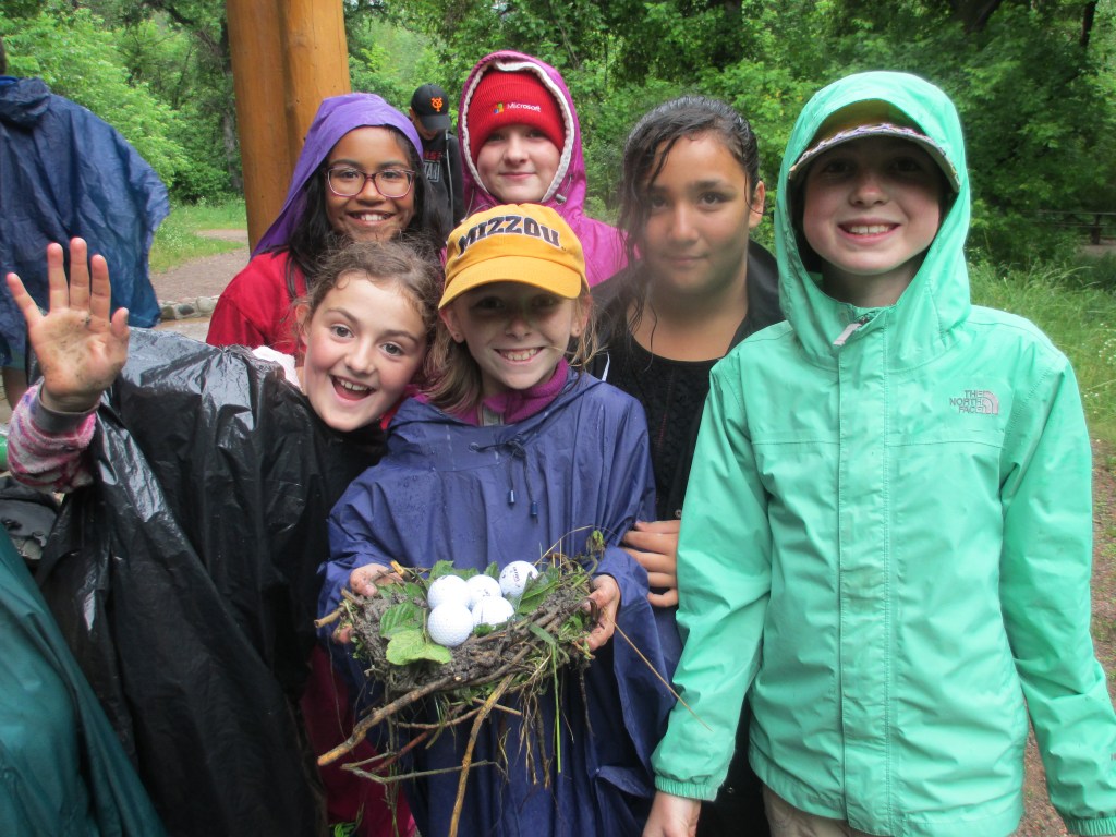 Six youth smile at the camera. In the center, a young girl holds a nest with eggs.