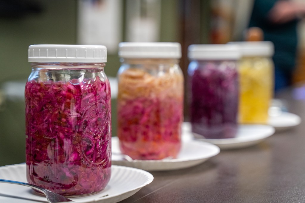 A line of four mason jars filled with different pickled vegetables, all atop paper plates. A spoon rests on the one closest to the camera.
