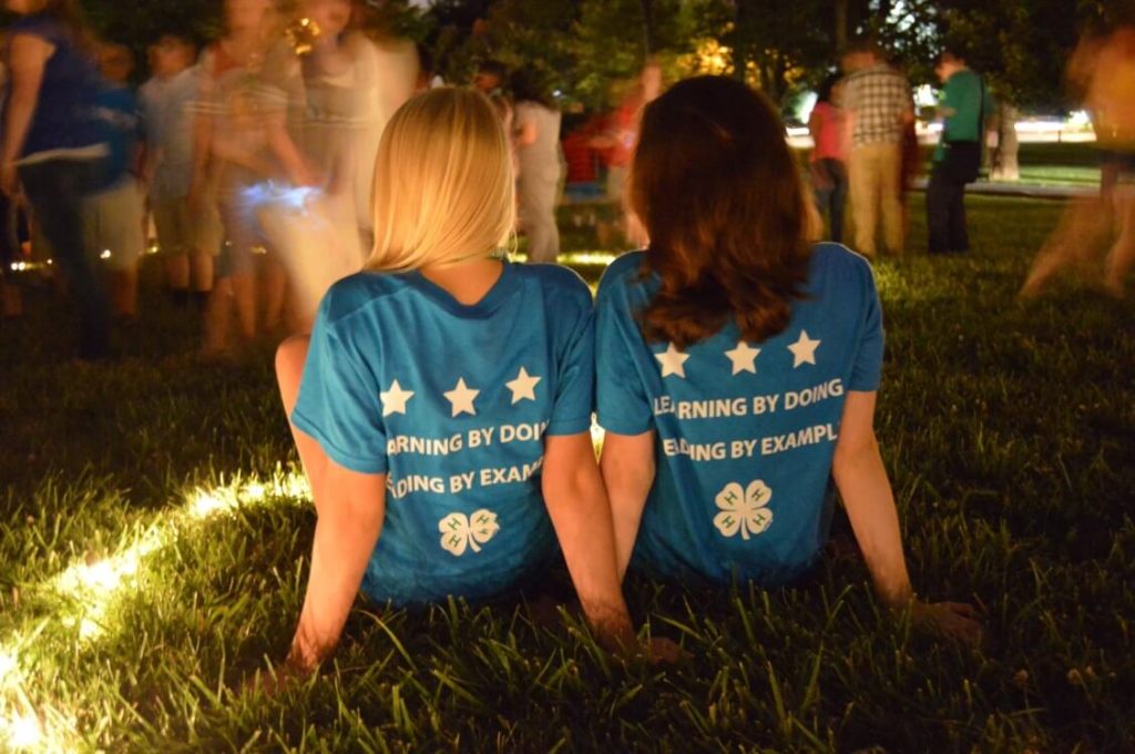 Two kids wearing matching blue 4-H shirts sit on the grass at night with their backs to the camera while people move around them in the background.