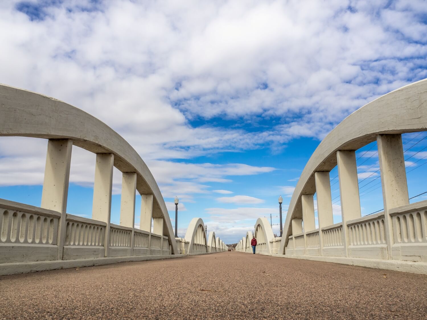 A wide-angle view of a concrete arch bridge with repeating curved supports, leading toward the horizon under a bright sky filled with scattered clouds.