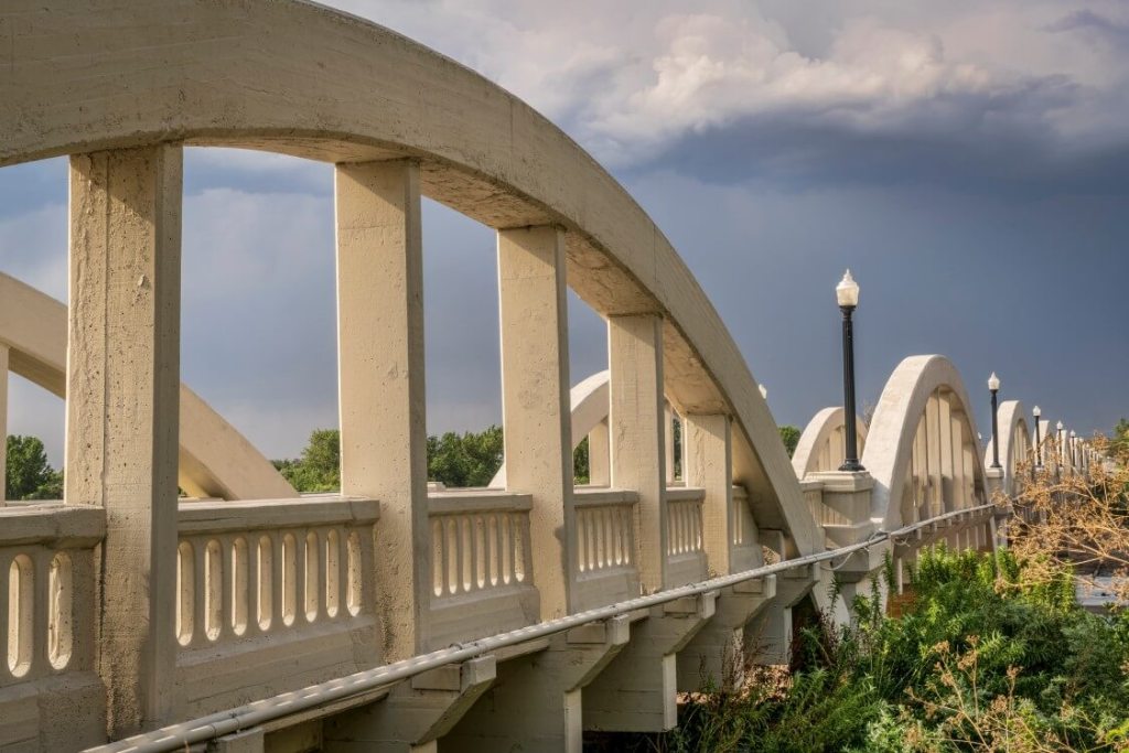 Side view of a concrete arch bridge with repeating curved supports and railing details, extending into the distance with streetlamps and trees below under a partly cloudy sky.
