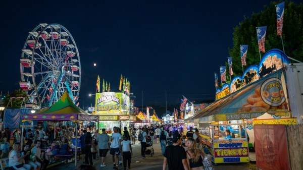 A lively nighttime county fair scene featuring a Ferris wheel, illuminated food booths, colorful banners, and crowds walking along a midway lined with carnival games and attractions.