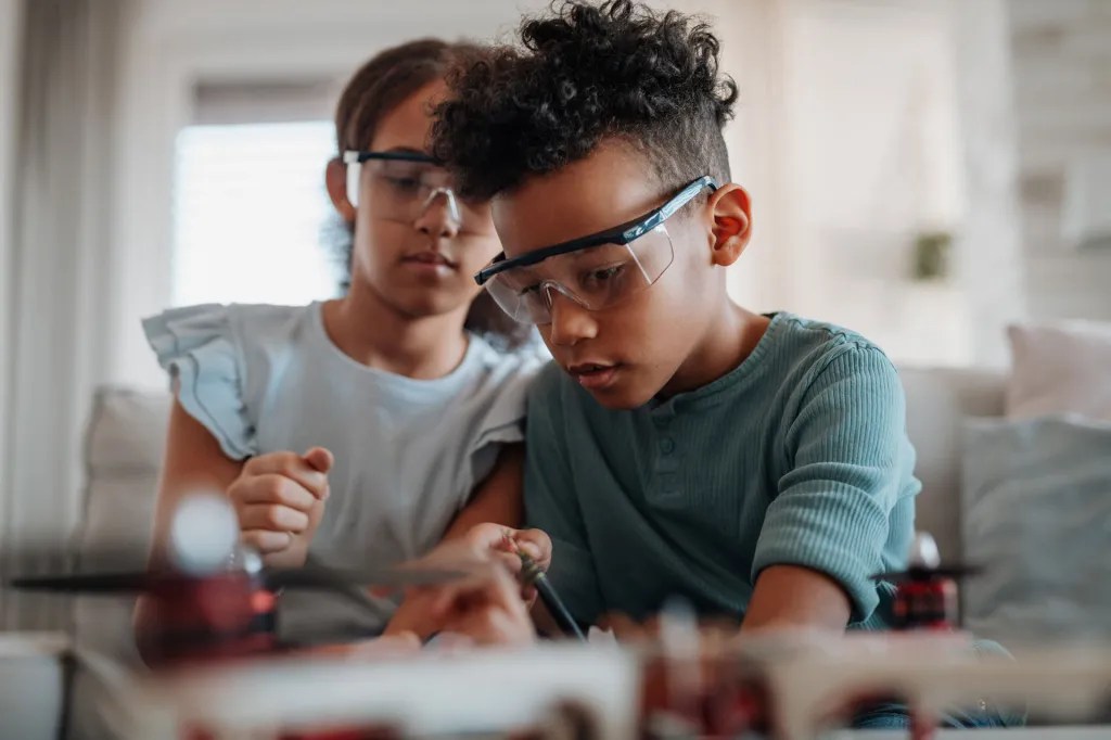 Two children sit close together indoors, operating a small drone or remote‑controlled device on a table in front of them.