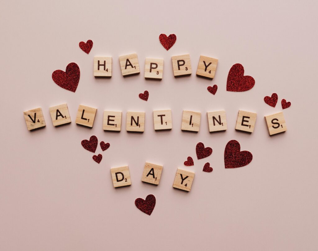 Scrabble tiles arranged to spell out "Happy Valentine's Day," surrounded by red glittery heart shapes on a light pink surface.