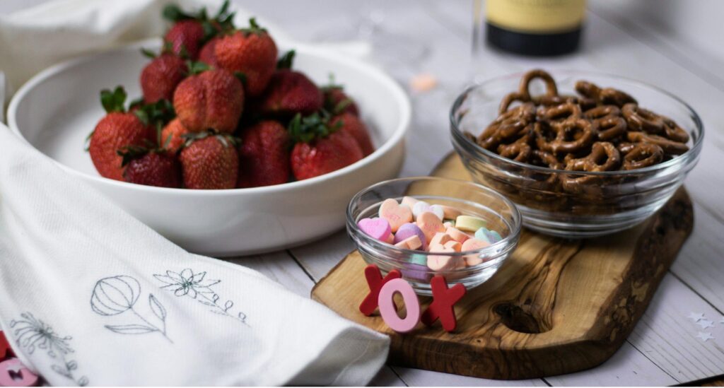 A wooden serving board with bowls of strawberries, chocolate-covered pretzels, and colorful heart-shaped candies.