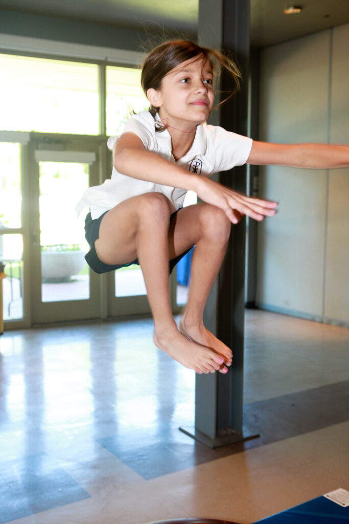 A young girl in a white polo shirt and black shorts jumps in the air indoors, appearing focused and determined.