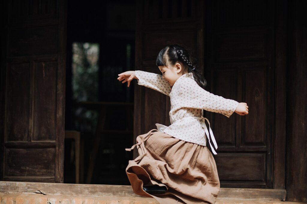 A young girl in a long skirt and white patterned blouse jumps off a wooden step outside a rustic building, arms spread wide.