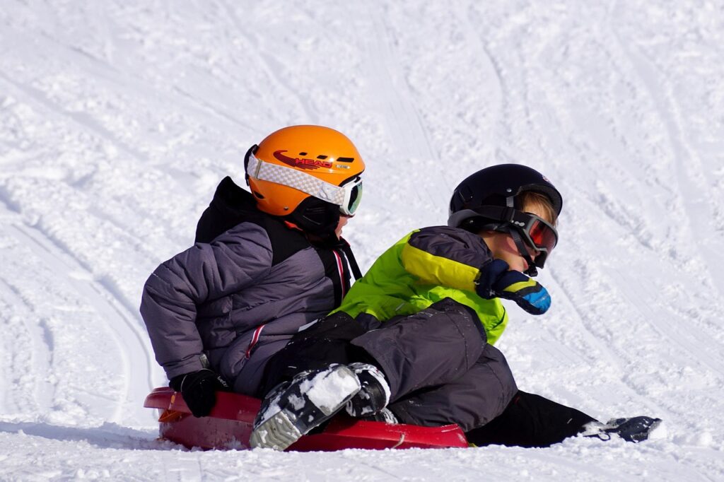 Two children wearing winter gear and helmets ride a red sled down a snowy hill, leaning into the turn.