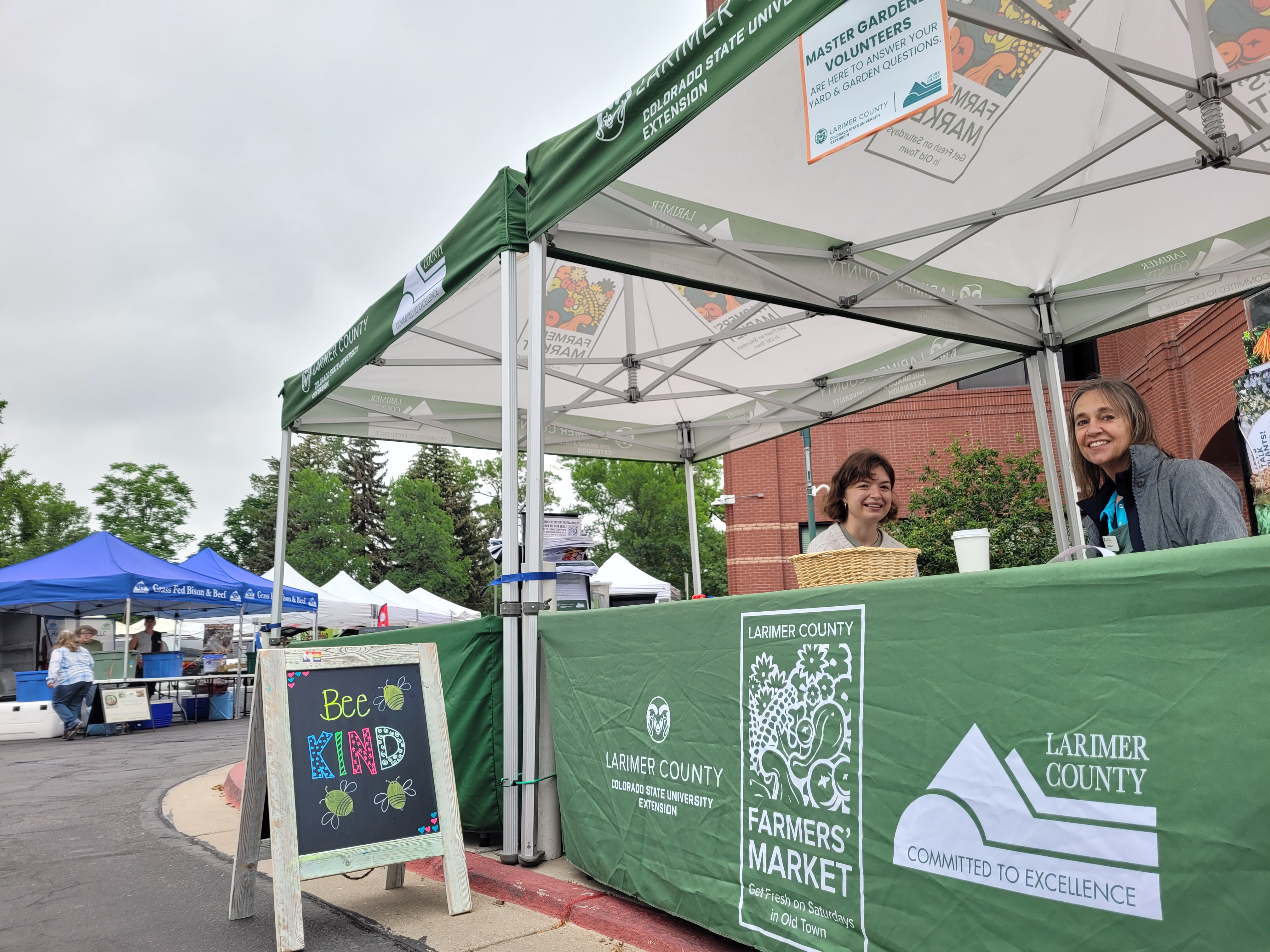 Two women smiling under a Larimer County Farmers’ Market tent, with a “Bee Kind” sign and other vendor booths in the background.