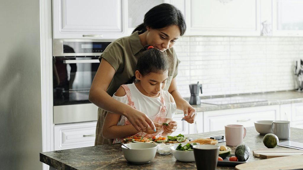 Mother and daughter preparing fresh sandwich with lettuce, avocado, tomatoes, etc in their kitchen.