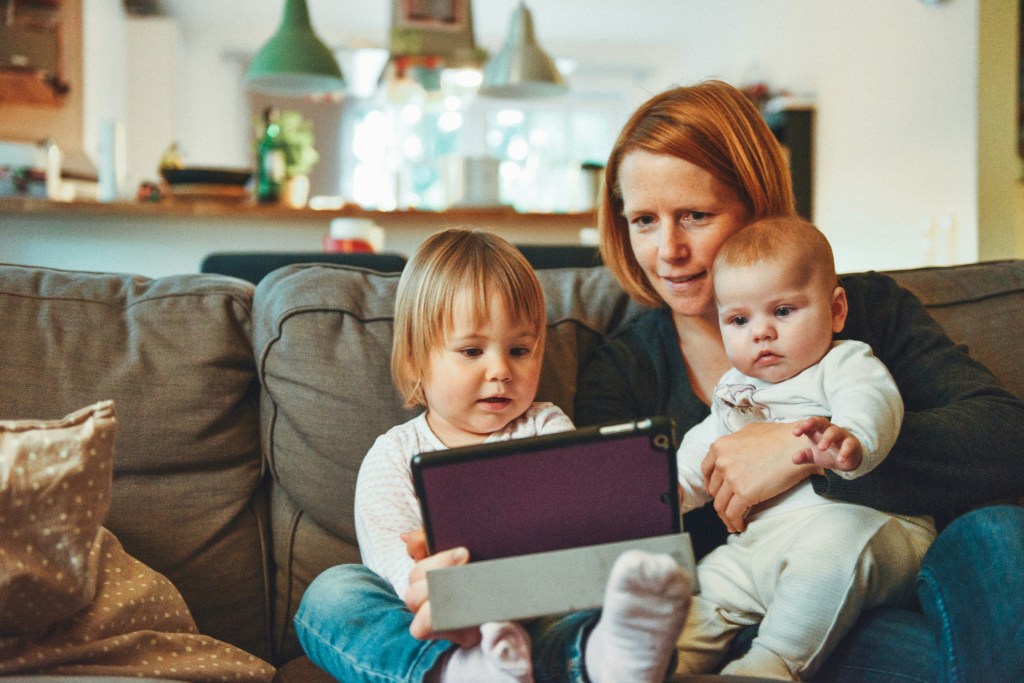 A mother sits on a couch with her two young children, watching something together on a tablet.