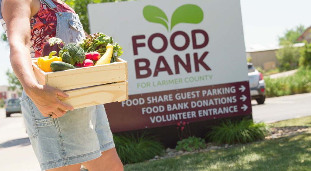 Woman in overall shorts holding a wooden box full of fresh vegetables including broccoli, squash, zucchini, bell peppers, with a Food Bank for Larimer County in the background.