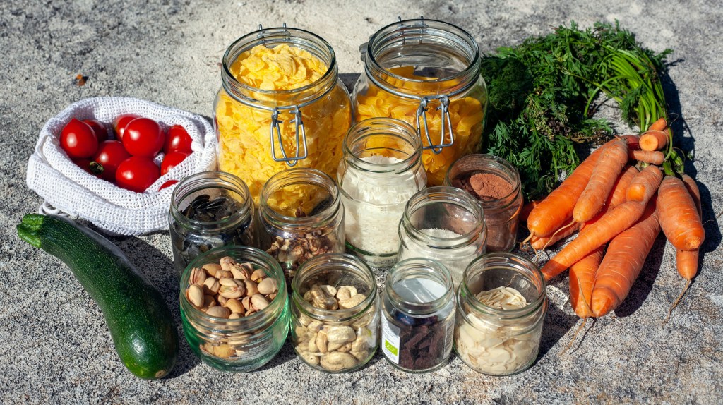 Top view of glass jars filled with nuts, seeds, and grains, surrounded by fresh vegetables like carrots, zucchini, and cherry tomatoes on a stone surface.