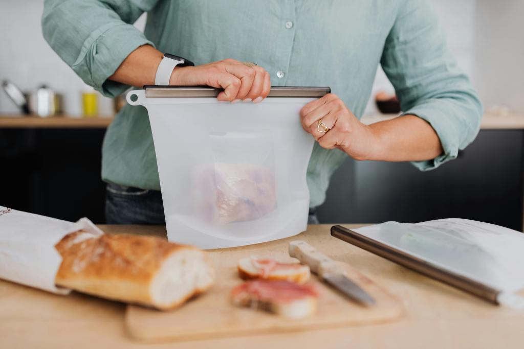 Person sealing a reusable silicone bag with a sandwich on a wooden kitchen counter, next to sliced baguette and a knife.