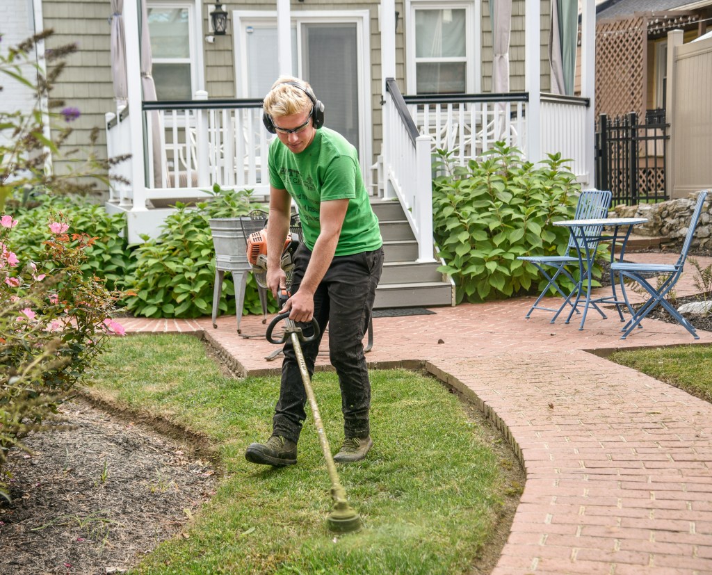 A person wearing ear and eye protection uses a string trimmer to edge the grass along a brick path in a landscaped backyard.