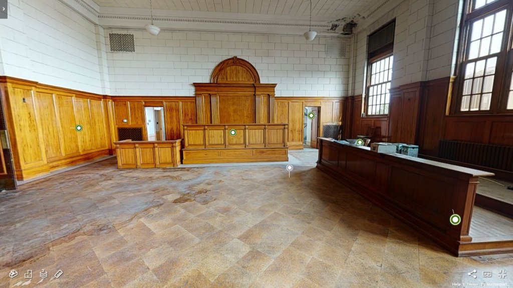 Empty courtroom with wood paneling, a judges bench, witness stand, and attorney table. Large windows on the right let in natural light with worn down tile flooring.
