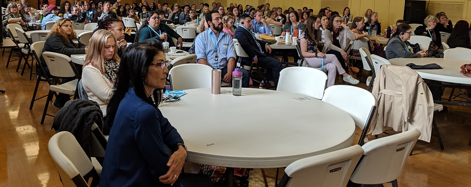 Large group of adults seated at round tables in a gymnasium or hall, attentively listening to a speaker out of frame during a conference or community event.