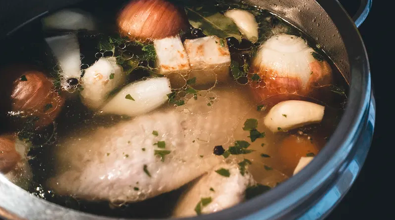 Closeup of chicken, onions, garlic, and fresh herbs in a broth cooking in a slow cooker. 