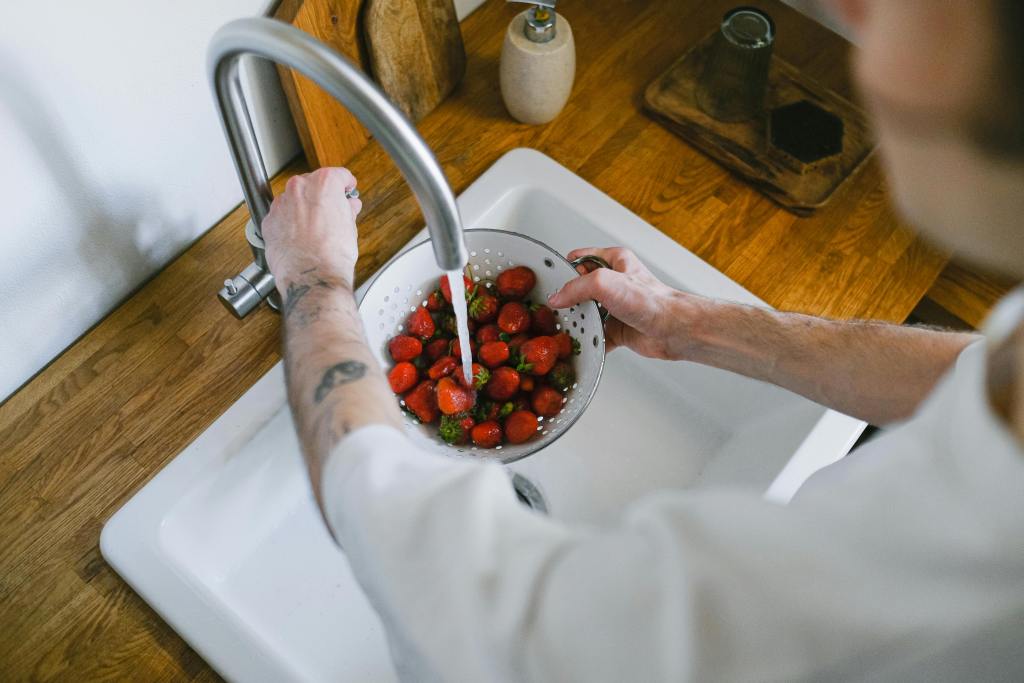 Person rinsing a colander full of fresh strawberries under running tap water in a kitchen sink.